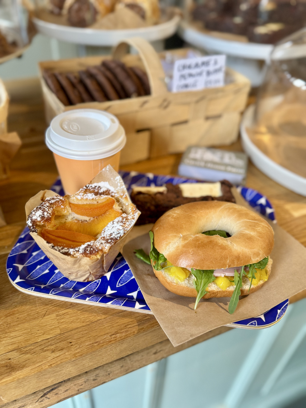 A bagel, muffin and coffee on a tray in front of a basket of chocolate cookies
