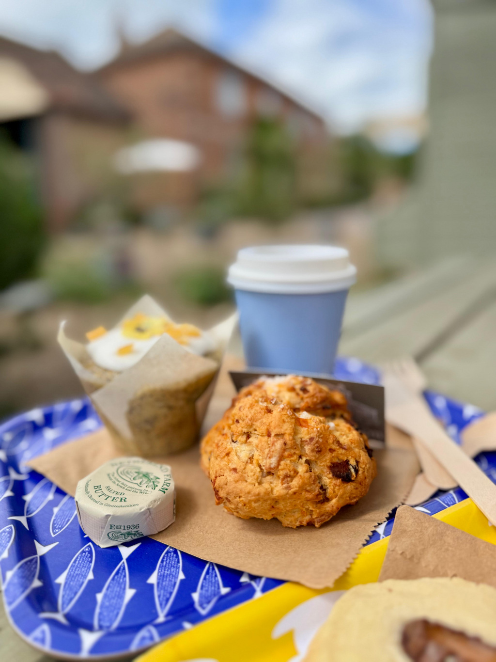 A scone, muffin, and coffee sitting on a tray with a wrapped square of butter, with a blurred house 