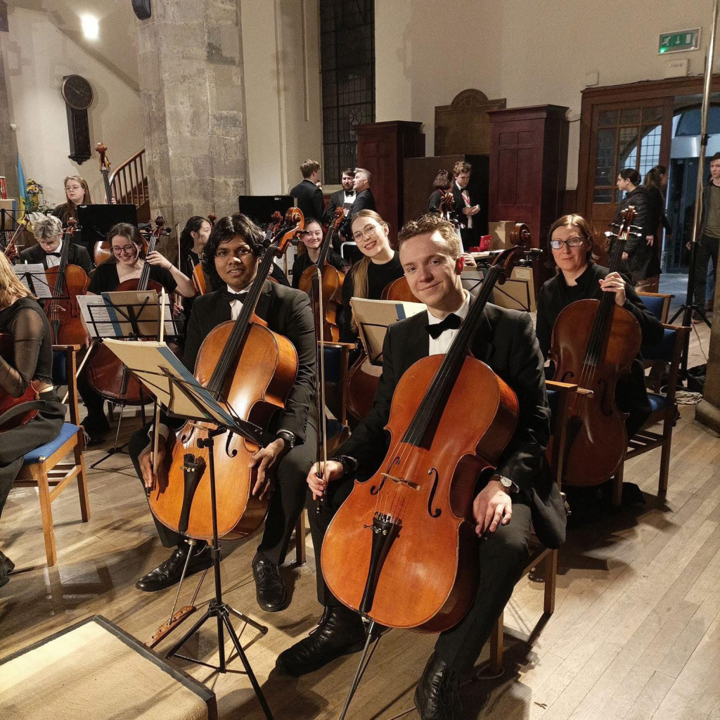 Two men sitting in an orchestra formation with cellos on their knees