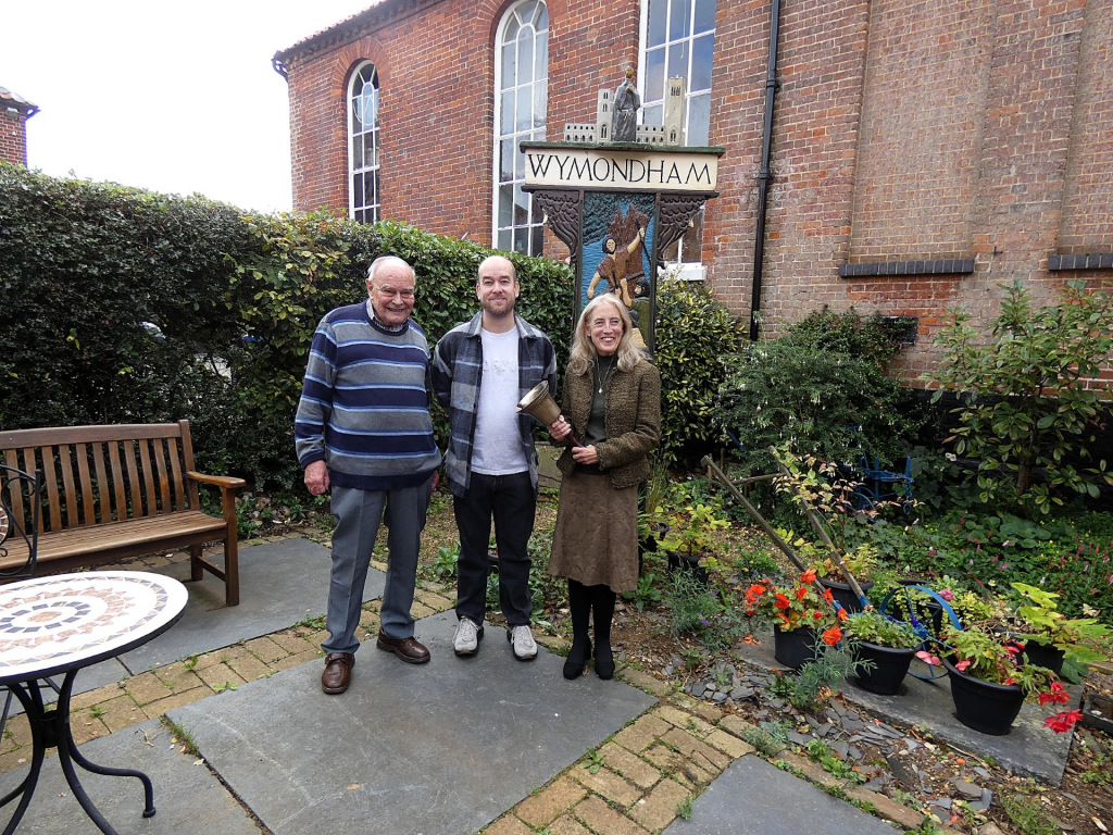 Two men and a woman standing by a 'Wymondham' sign in a garden