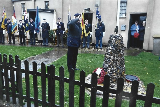 An officer in a garden saluting a monument surrounded by a brass band