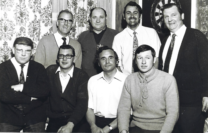 A black-and-white still of a group of men with a dartboard behind them