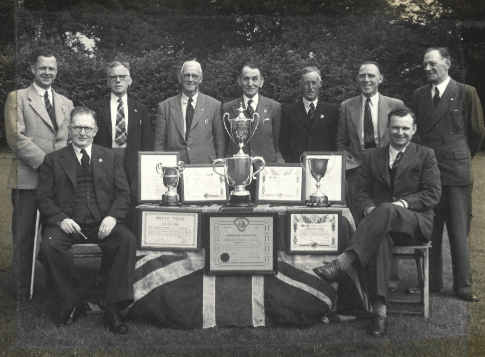 Trophies and certificates on a table surrounded by gentlemen in suits